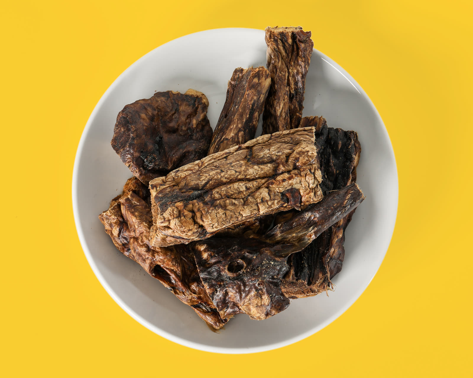 Water buffalo lung treats in a white bowl, shot overhead on a yellow seamless background